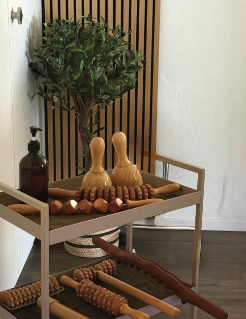 Beistellwagen mit Holzwerkzeugen für die Maderotherapie — Rollen, Glocken und ein Brett — vor einer Holzlattenwand.