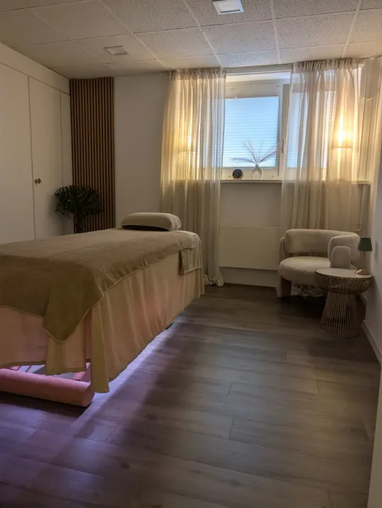 Behandlungsraum mit bezogener Massageliege, einem Sessel am Fenster und Tageslicht hinter leichten Vorhängen.